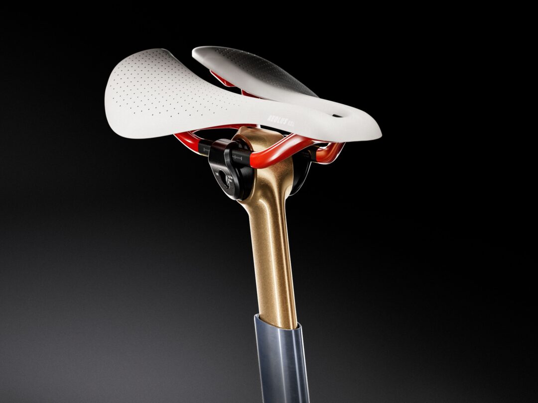 Close-up of a white perforated bicycle saddle with red rails mounted on a bronze seatpost against a black gradient background.