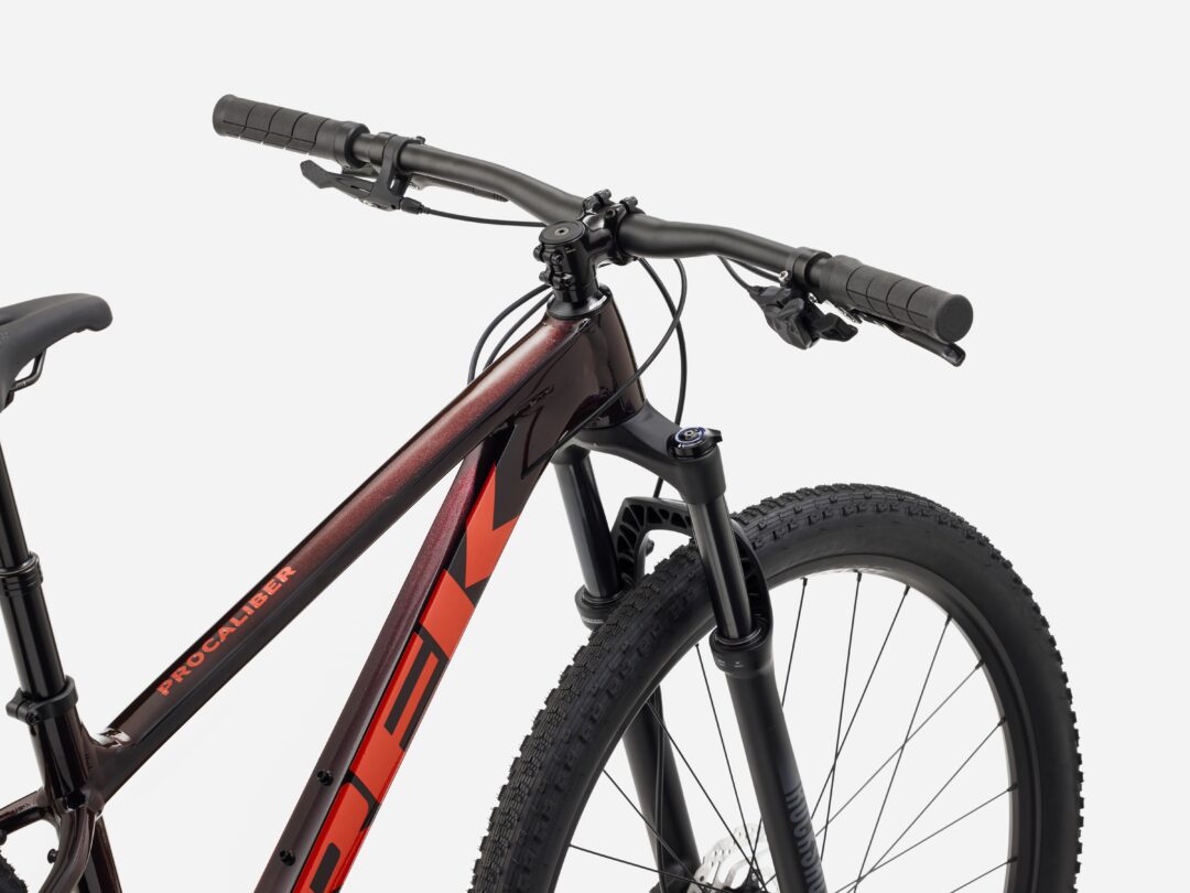 Close-up of a dark red mountain bike showing the handlebar, grips, brake levers, cables, and front suspension fork with a large wheel partially visible.