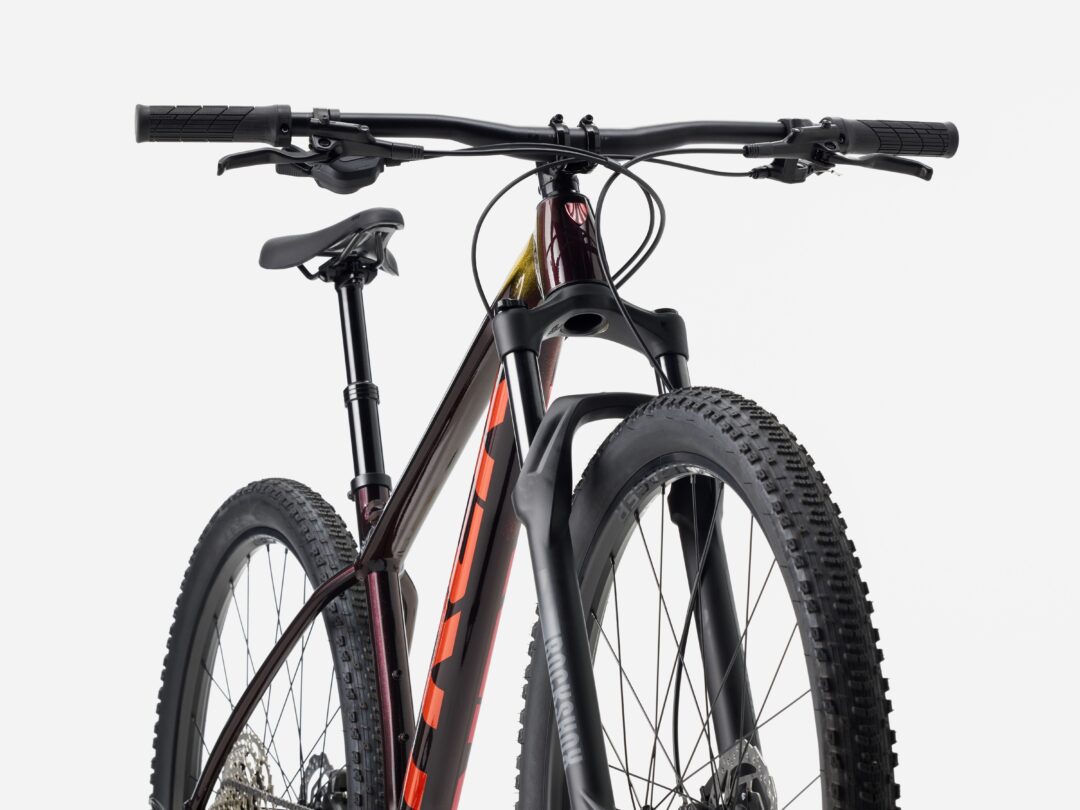 Front view of a mountain bike cockpit with wide flat handlebars, cables, and knobby tires visible around the front wheel. Frame colors include maroon and orange accents.